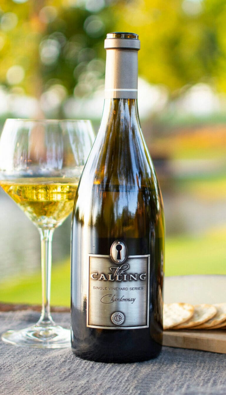 Bottle of 2018 Sullivan Chardonnay resting on table, with crackers on its right and glass of wine on its left.