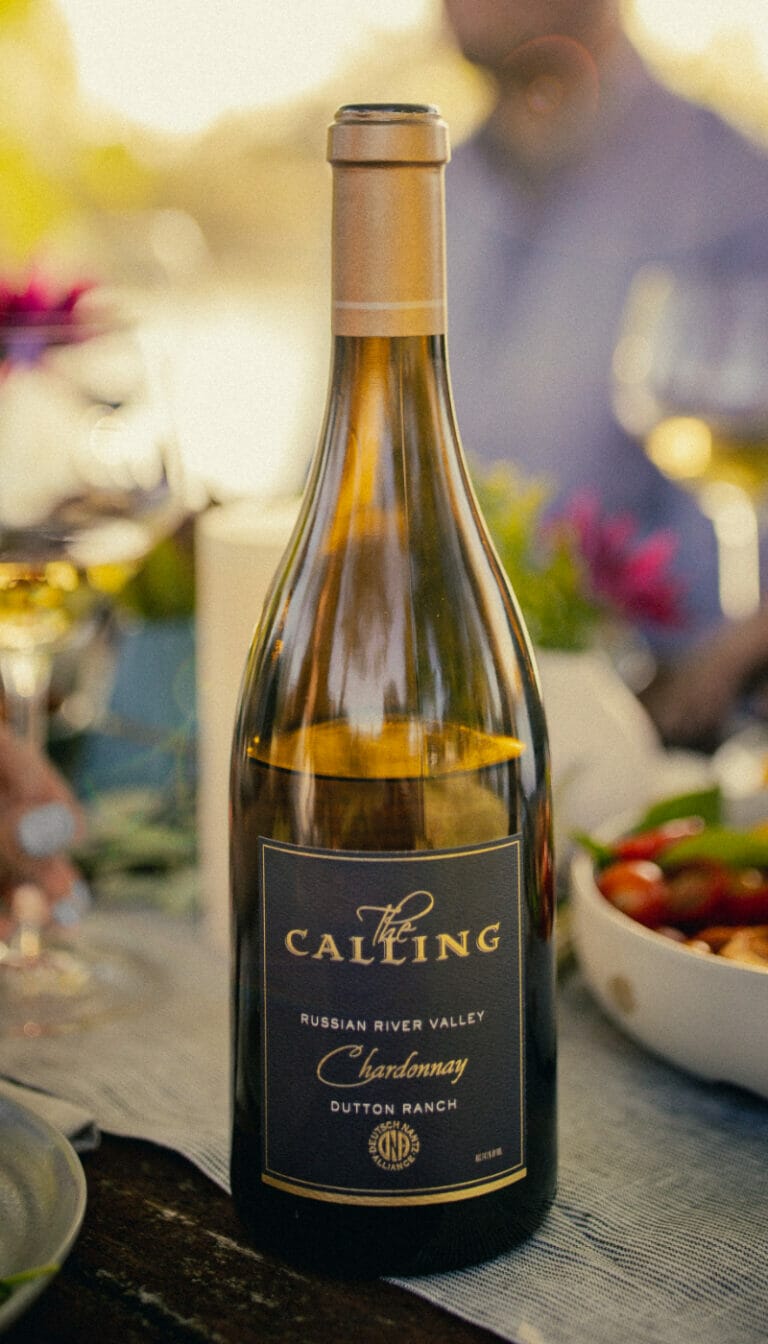 Bottle of 2019 Dutton Ranch Chardonnay displayed on table, with people and food in background.