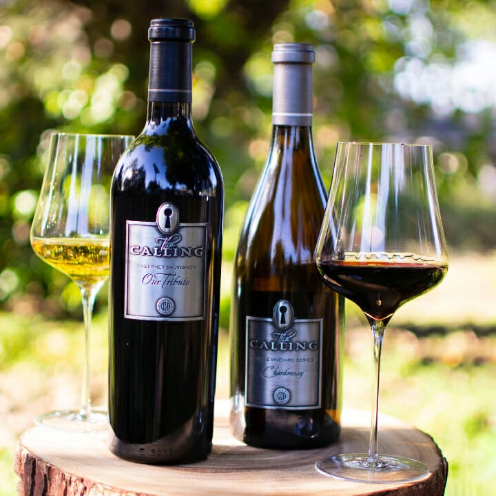 Two bottles of The Calling Wines resting on tree stump, with a glass of Chardonnay on one side and glass of Cabernet Sauvignon on the other.
